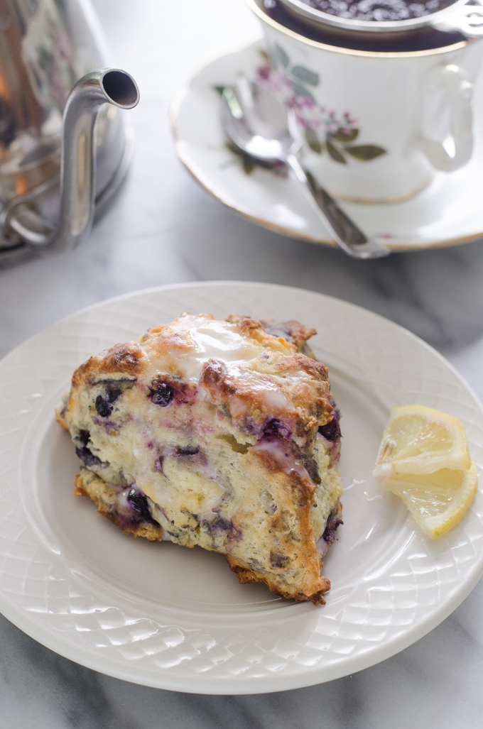 Blueberry scones on a dessert plate with small slices of lemon off to the side and a cup of tea and a tea pot in the background.