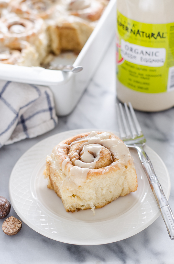 An individual eggnog cinnamon roll on a plate with the pan in the background.