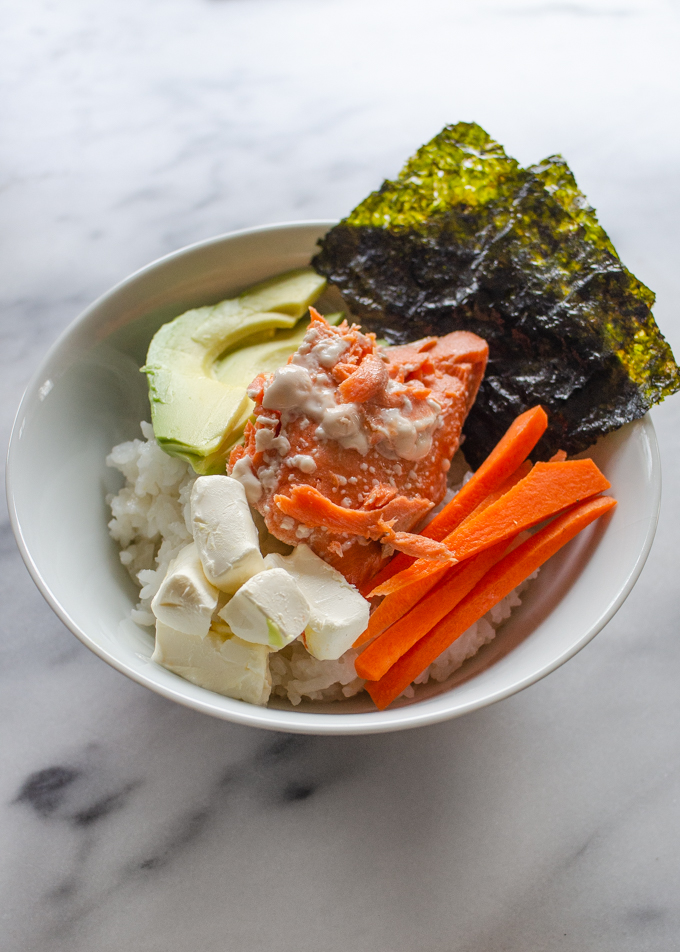 A bowl of sushi rice topped with avocado, seaweed snacks, carrots, cream cheese, and salmon.