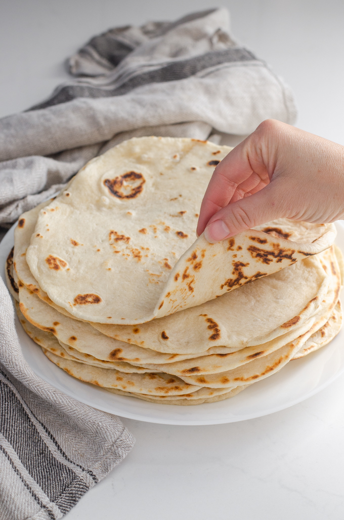 Sourdough tortillas stacked on a plate with a tea towel around them with a hand rolling the top tortilla.