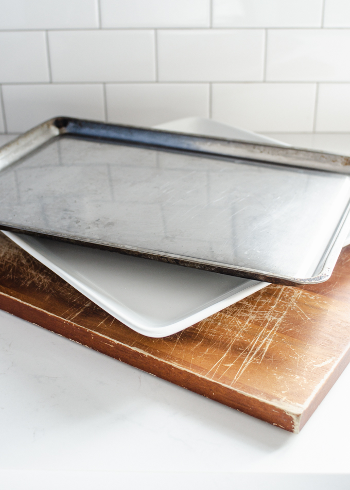 A cutting board, large plate, and cookie sheet stacked on top of each other.