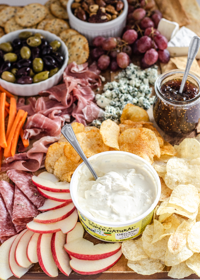 A snack board with nuts, crackers, grapes, brie cheese, fig spread, olives, blue cheese, prosciutto, carrots, chips, apples, salami, and Kalona SuperNatural French onion dip.