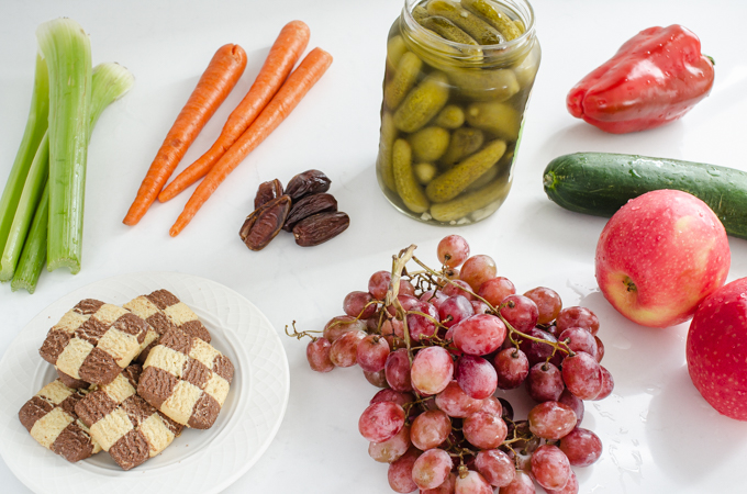 Fresh fruits and veggies, pickles, and cookies laid out on a counter. 