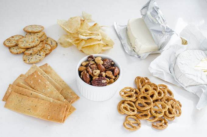 Crackers, chips, roasted nuts, pretzels, cream cheese, and brie cheese laid out on a counter.