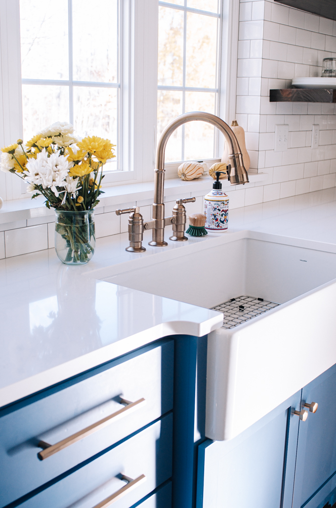 White apron front sink in front of window with bronze/gold bridge faucet in a transitional modern farmhouse kitchen.